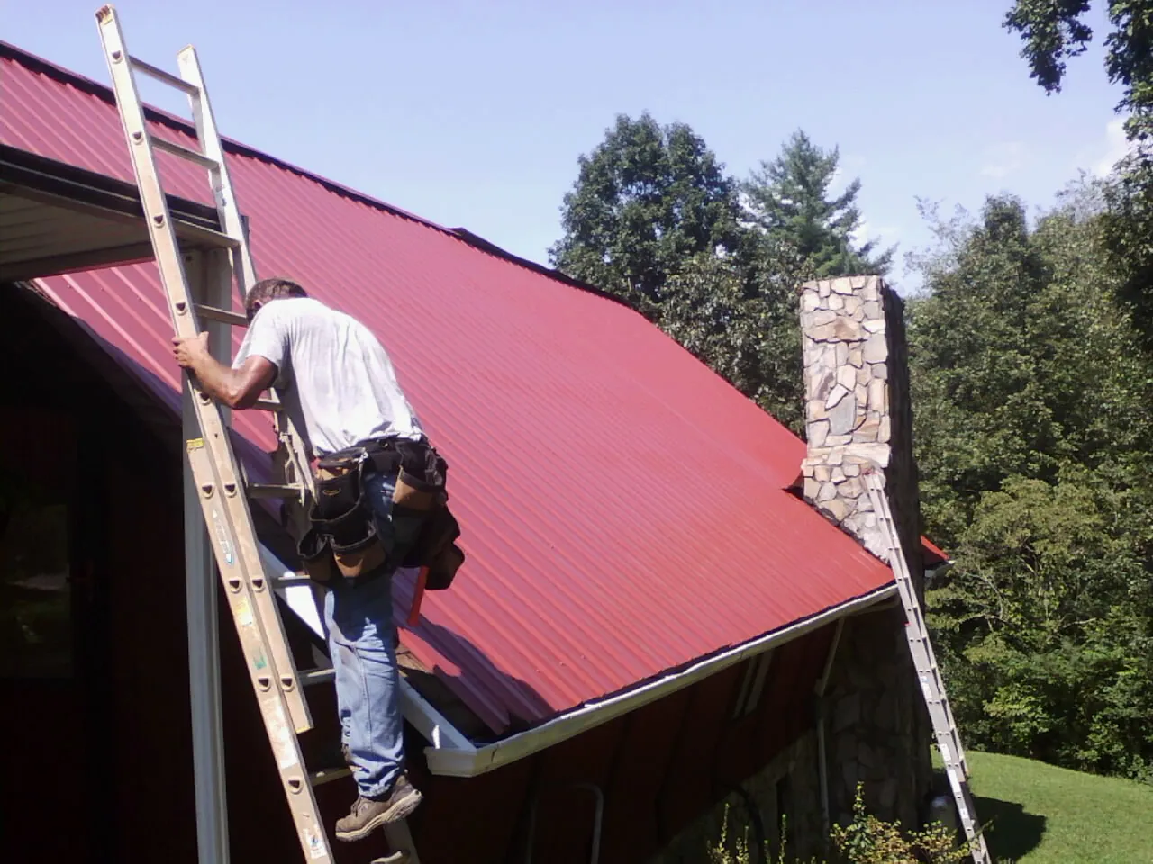 Roofer on a ladder beside a red metal roof and stone chimney — Soffit & Fascia Repair by Roofing Brothers in Dendron
